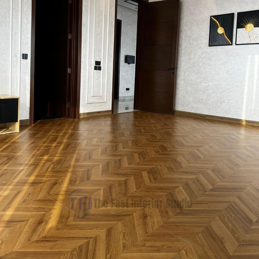 A bright, sunlit room featuring elegant Chaina Floor 12 MM chevron laminate flooring with a warm golden amber finish, seamlessly installed next to a dark wood door