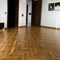 A bright, sunlit room featuring elegant Chaina Floor 12 MM chevron laminate flooring with a warm golden amber finish, seamlessly installed next to a dark wood door