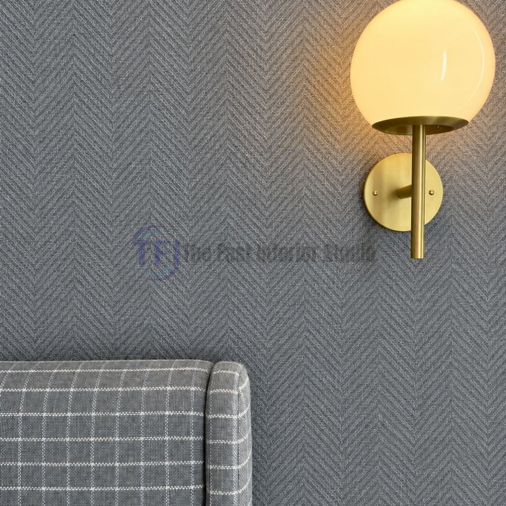 A close-up interior shot of a gray herringbone textured jute wallpaper installed on a bedroom accent wall, styled next to a modern brass globe wall sconce and a gray checkered upholstered headboard