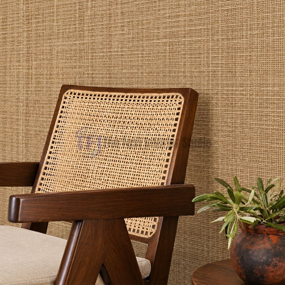 A close-up interior shot of a natural golden-brown jute wallpaper accent wall styled behind a wooden armchair with a cane-back weave and a potted plant