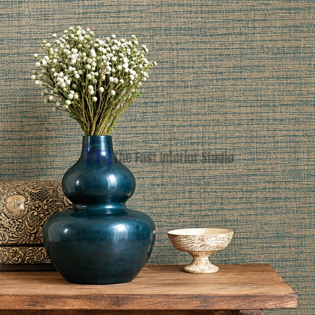 A close-up interior shot of a rich teal and brown woven jute textured wallpaper installed behind a wooden console table styled with a large glossy teal vase, white floral stems, and a decorative box