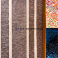 A close-up interior shot of dark brown jute wallpaper texture installed inside vertical white wainscoting wall paneling, displayed next to a framed colorful and blue calligraphic art piece
