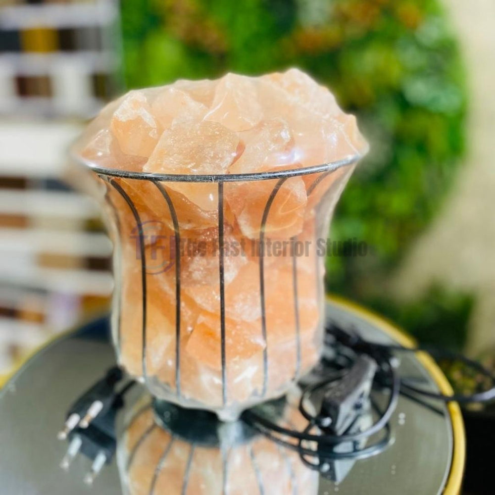 A contemporary Himalayan salt lamp featuring raw pink salt chunks inside a flared metal wire basket, emitting a warm amber glow