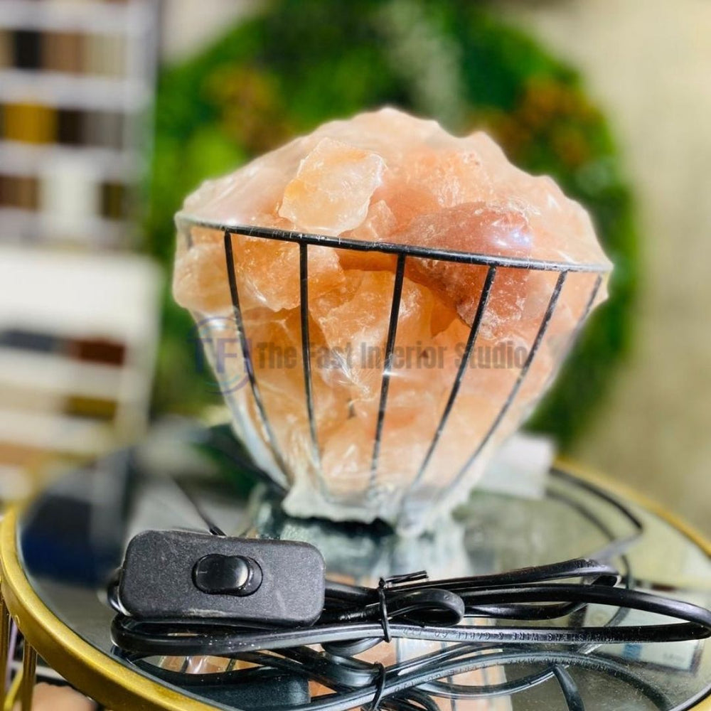 A contemporary Himalayan salt lamp featuring raw salt crystals displayed inside a flared black metal wire basket, providing a warm amber glow