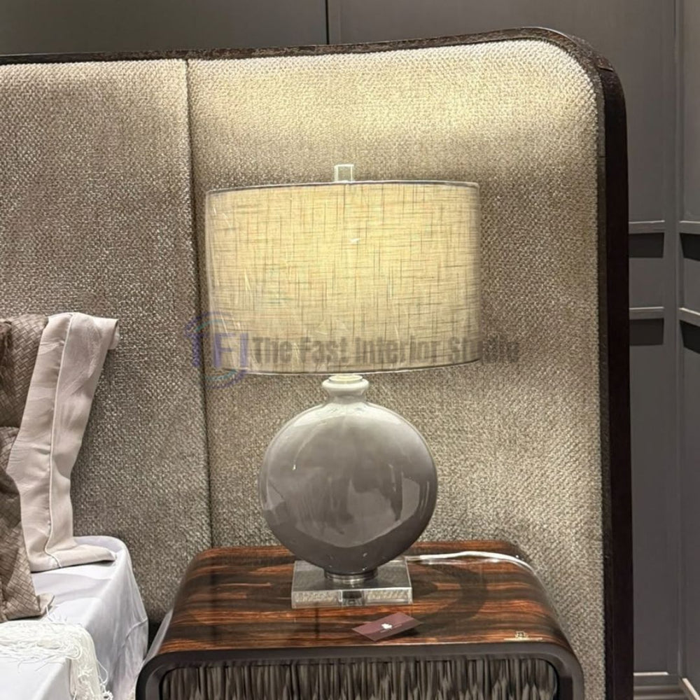 A contemporary table lamp featuring a dove grey ceramic orb on a crystal base with a textured linen shade, styled on a high-gloss wooden nightstand