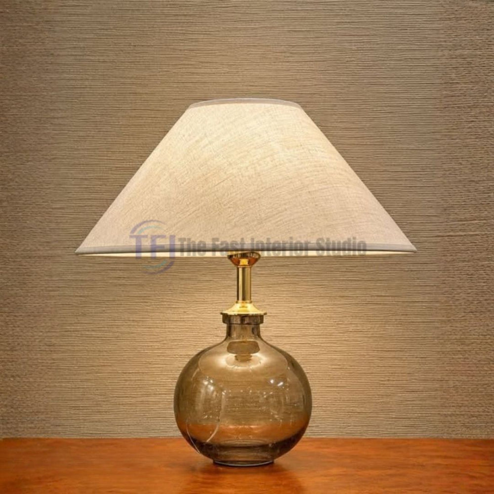 A contemporary table lamp featuring a round smoky glass base, gold brass neck, and a wide beige linen shade, styled on a wooden table for luxury interiors