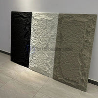 A display of three large vertical 3D PU chiseled faux stone wall panels in black, white, and warm beige, resting against a white wall in a modern setting