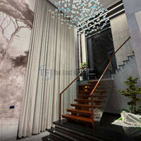 A grand foyer interior featuring extra-long, floor-to-ceiling neutral curtains installed beside a wooden staircase with glass railings, a large landscape mural, and a blue glass butterfly chandelier