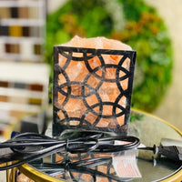 A minimalist Himalayan salt lamp featuring raw pink salt chunks displayed inside a cylindrical black metal wire basket with vertical bars