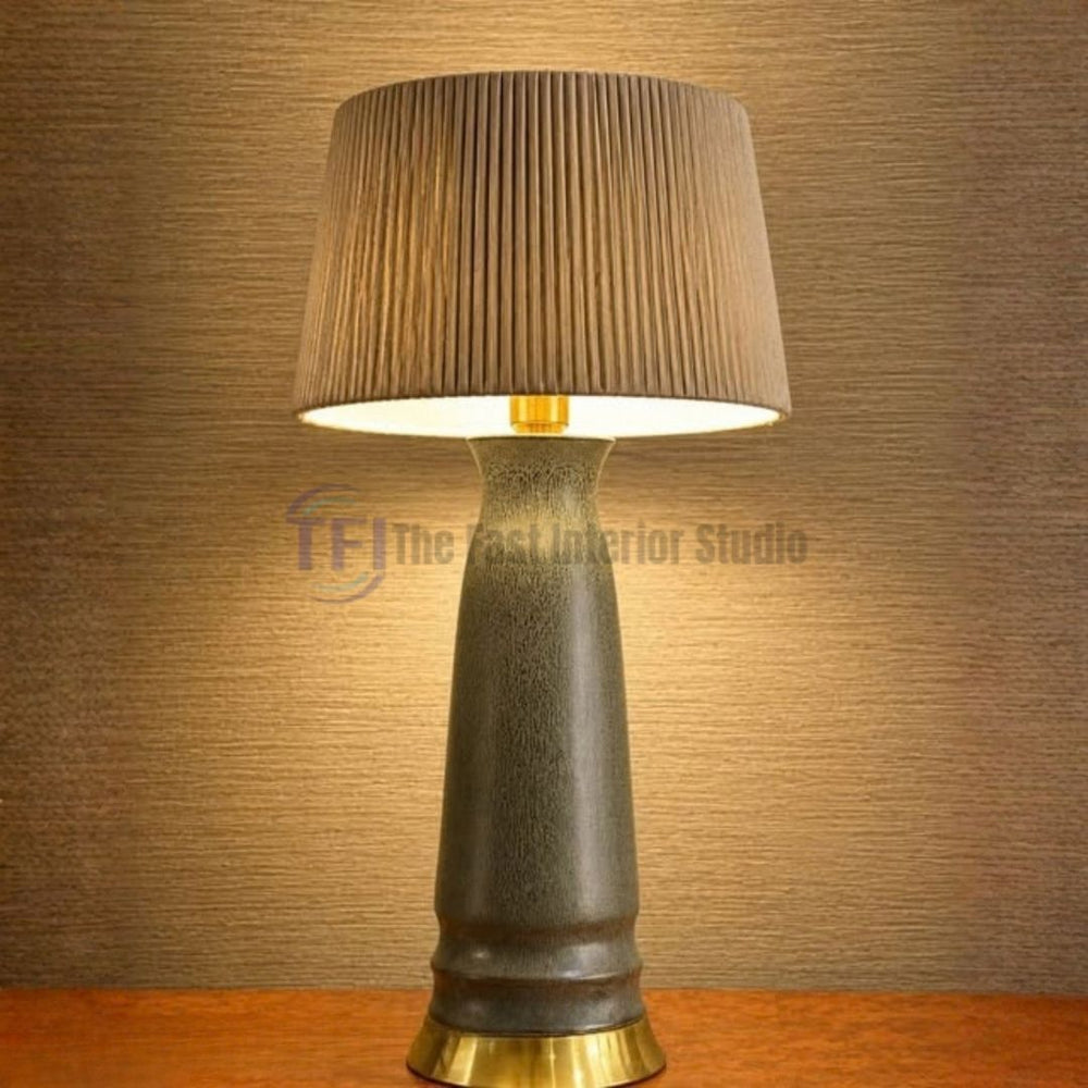 A premium textured charcoal ceramic table lamp featuring a gold brass base and a taupe pleated shade, styled on a wooden surface for luxury interiors