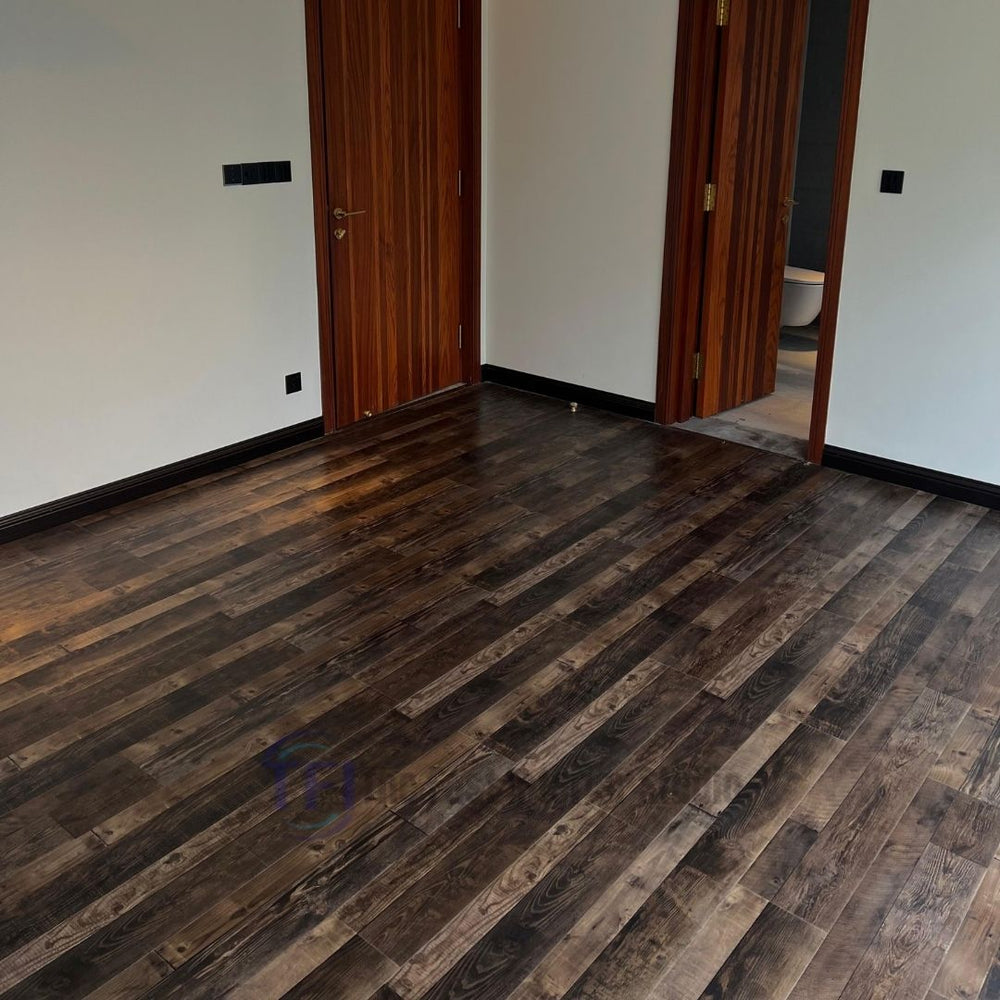 An empty room showcasing expansive multi-tone rustic Chaina Floor 12 MM laminate flooring, providing a bold architectural base against crisp white walls and dark ribbed doors