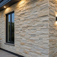 An exterior property wall cladded with light beige 3D PU stacked faux stone panels, beautifully illuminated by modern architectural sconce lighting