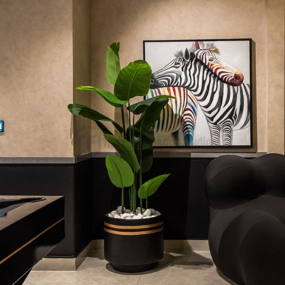 Tall 6-foot artificial plant with broad green leaves in a black pot with gold stripes next to a zebra painting