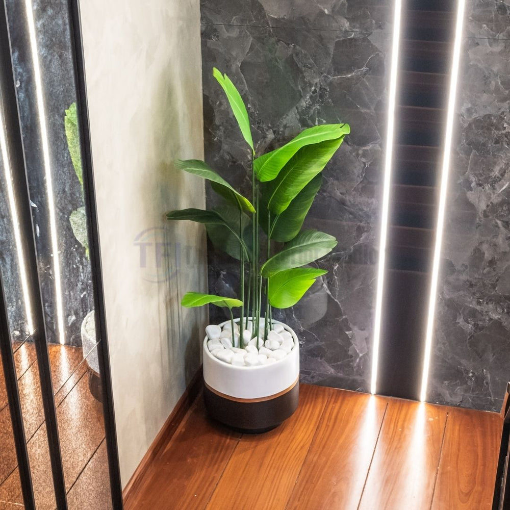 Tall 6-foot artificial plant with broad green leaves in a two-tone white and black pot filled with white stones on a wood floor