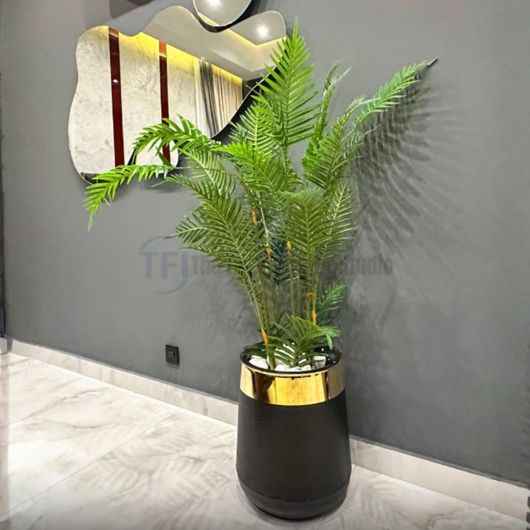 Tall artificial fern plant in a black pot with a gold rim filled with white stones against a grey wall
